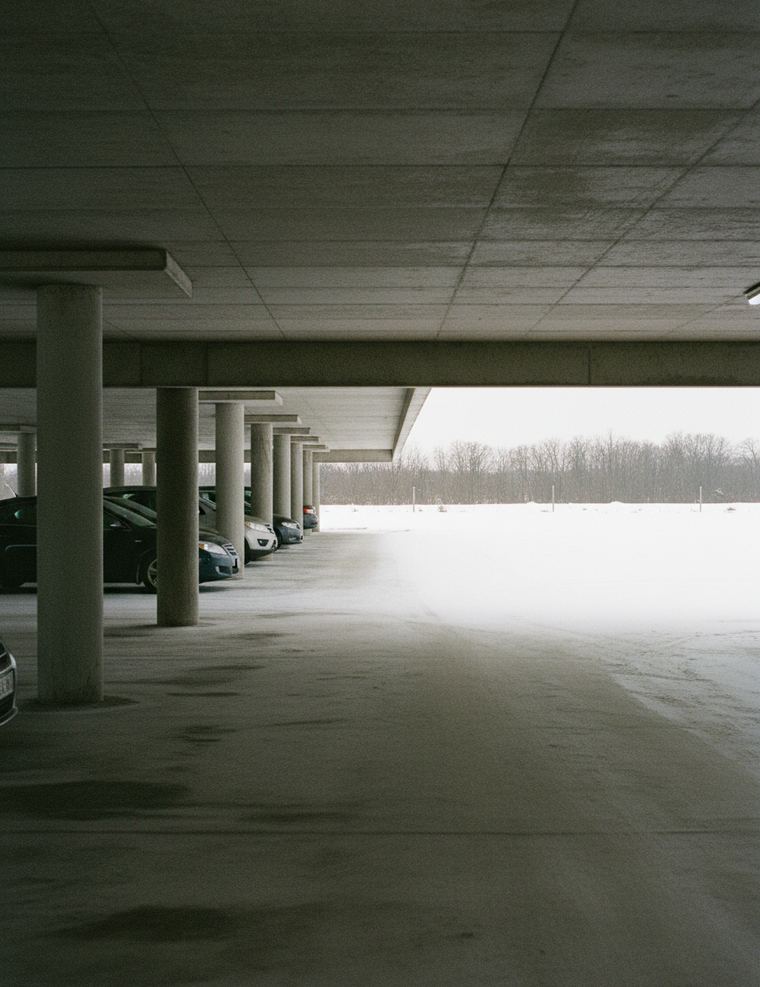 The lower level of a parking garage with a concrete ceiling and round columns, a few cars parked inside and a flat snowy field beyond the open side
